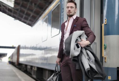 Train Leaders - Serious stylish bearded businessman in trendy suit holding bag and coat in hands standing near train on platform in railway station and looking away