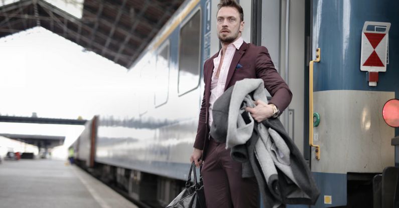 Train Leaders - Serious stylish bearded businessman in trendy suit holding bag and coat in hands standing near train on platform in railway station and looking away