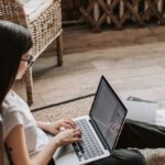 Sustainable Program - Young barefoot woman using laptop on floor near books in stylish living room