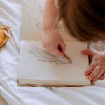 Small Businesses - From above of small girl in dress drawing with pencil in notebook while mother using laptop on bed near