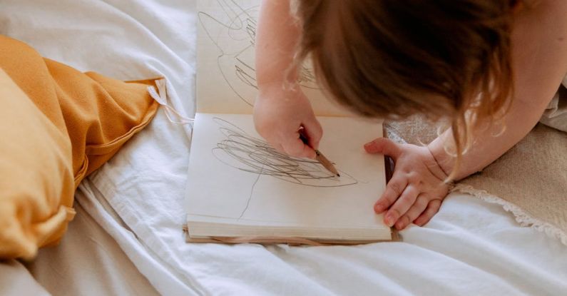 Small Businesses - From above of small girl in dress drawing with pencil in notebook while mother using laptop on bed near