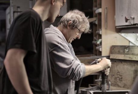 Legacy - Side view of senior artisan in eyeglasses handling detail in workshop while apprentice looking at process